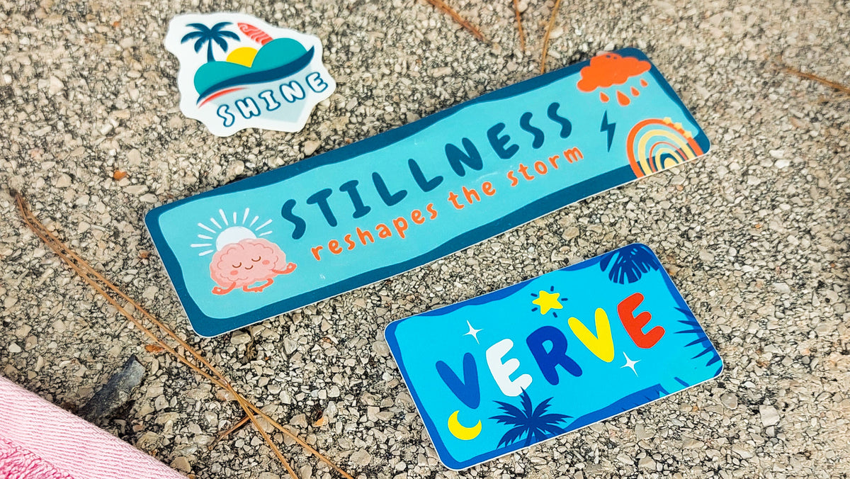Three colorful rectangular stickers with tropical and beach-themed designs placed on a gravel surface.